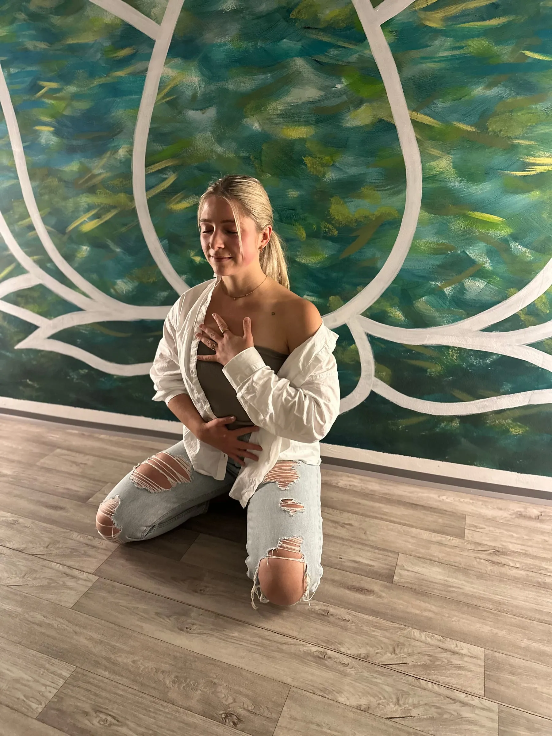 Katerina Kaslova sitting with her eyes closed, one hand on her heart and one on her belly, in front of a painted teal lotus mural