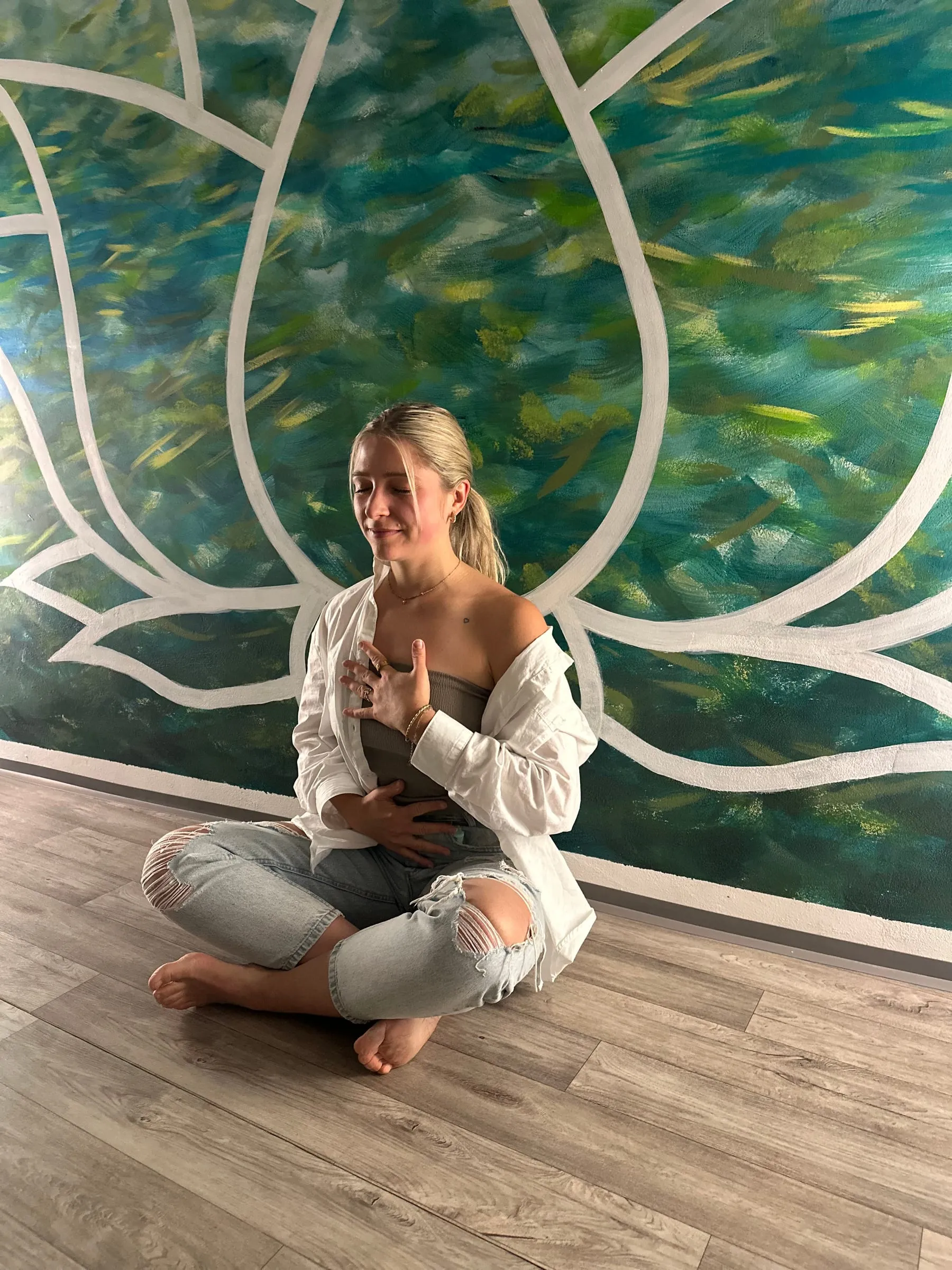 Katerina Kaslova sitting cross-legged and barefoot with her hands on her heart and belly, in front of a painted lotus mural