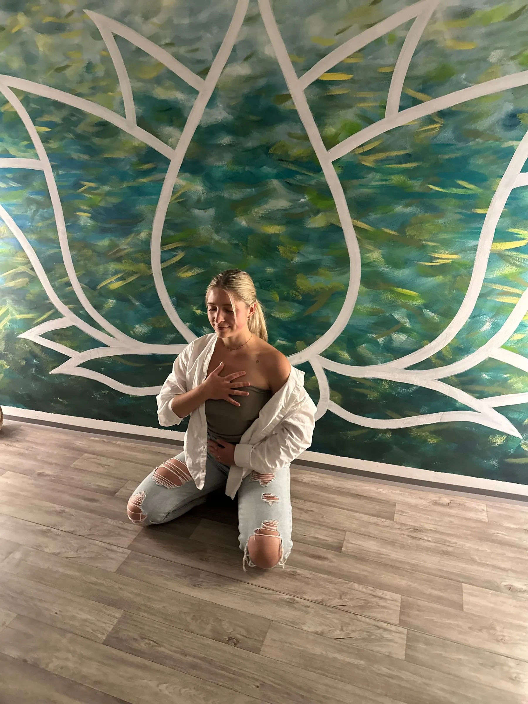 Katerina Kaslova kneeling with one hand on her heart and one on her belly, in front of a painted green and teal lotus mural